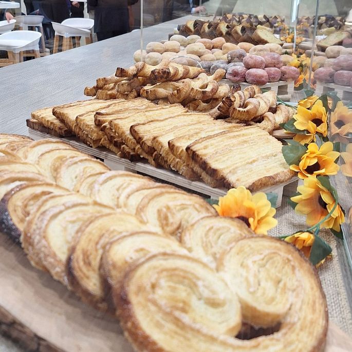 Close‑up of assorted pastries including palmiers, croissants and sugar‑coated doughnuts arranged on wooden boards behind a protective glass display with decorative yellow flowers.
