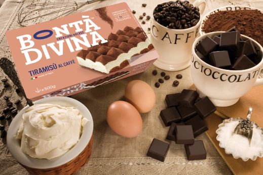Bontà Divina tiramisu box arranged with chocolate pieces, coffee beans, cocoa powder, eggs and whipped cream on a rustic surface.