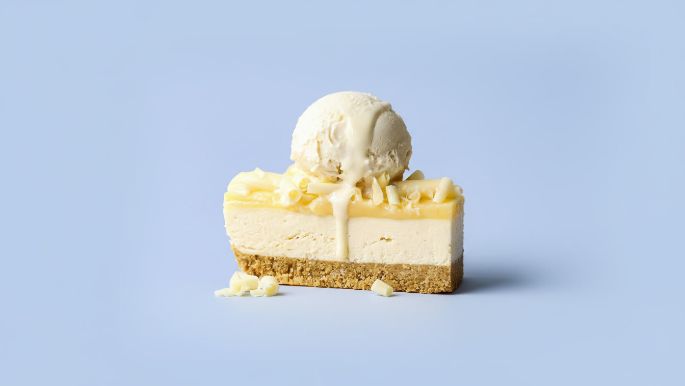 Slice of cheesecake topped with a scoop of vanilla ice cream and white chocolate shavings on a light blue background.