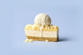 Slice of cheesecake topped with a scoop of vanilla ice cream and white chocolate shavings on a light blue background.