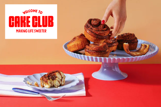 Cinnamon rolls on a cake stand with one piece on a plate, shown alongside the Cake Club logo.