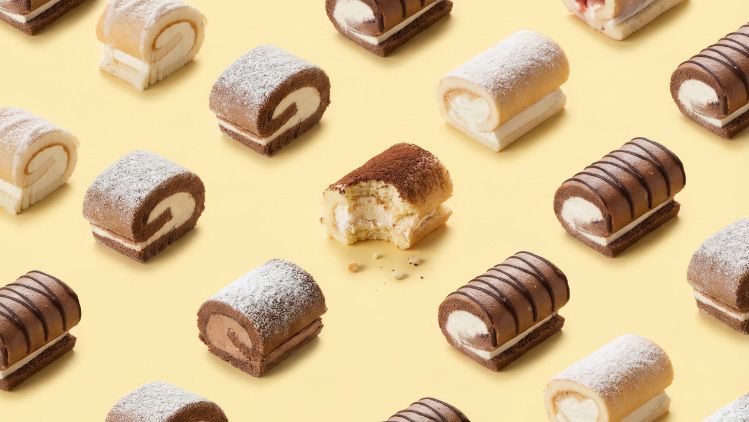 Assorted mini roll cakes in various flavors arranged in a repeating pattern on a yellow background, with one roll cut open to show the filling.