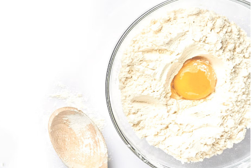 Top view of a bowl filled with flour and a cracked egg in the center, with a wooden spoon resting beside it on a white surface.