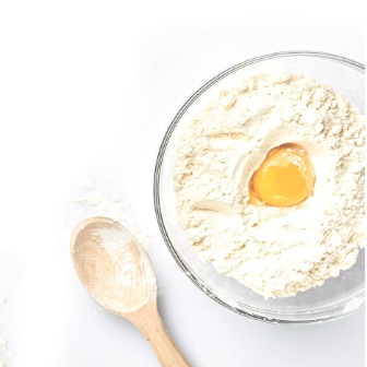 Top view of a bowl filled with flour and a cracked egg in the center, with a wooden spoon resting beside it on a white surface.