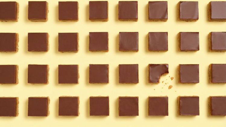 Grid of chocolate‑topped caramel shortbread squares on a yellow background, with one bitten piece showing crumbs