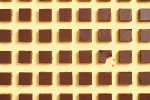 Grid of chocolate‑topped caramel shortbread squares on a yellow background, with one bitten piece showing crumbs