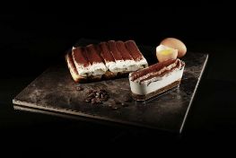 Tiramisu loaf and a sliced portion arranged on a dark stone slab, with cocoa powder, coffee beans and a cracked egg in the background.