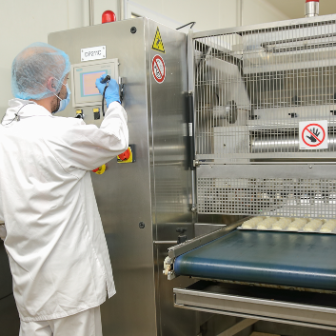 Person wearing protective clothing and a hair covering operating a stainless‑steel control panel next to food production equipment with a conveyor belt.