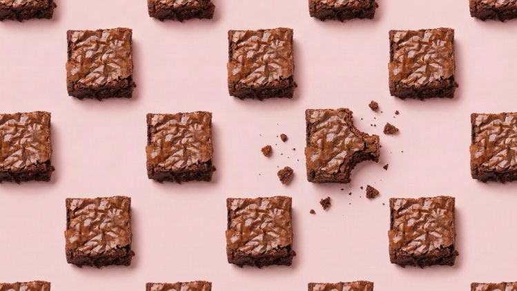 Pattern of chocolate brownies on a pink background with one bitten piece and scattered crumbs.