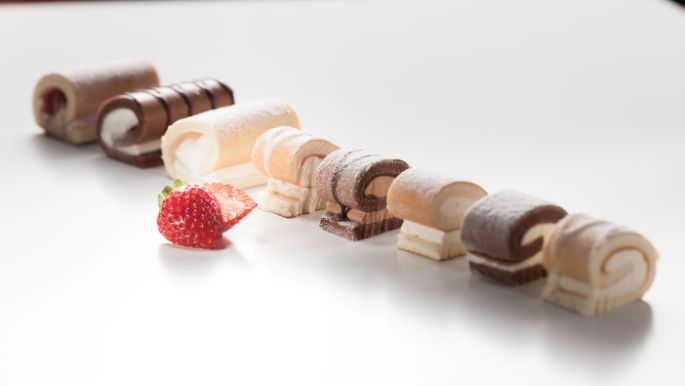 Assorted mini roll slices in various flavors arranged in a line with a fresh strawberry on a white surface