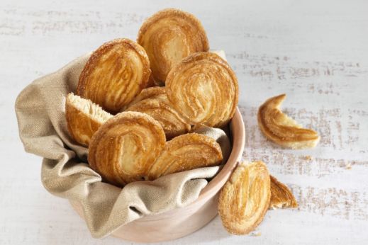 Bowl lined with a beige cloth filled with crispy palmier cookies, with a few additional palmiers placed on a light wooden surface.