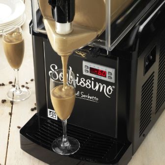 Sorbissimo machine dispensing a creamy chilled beverage into a glass, with additional filled glasses and scattered coffee beans on a wooden surface.