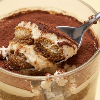 Close‑up of a spoon lifting a portion of tiramisu from a glass, showing layers of soaked biscuits, cream and cocoa powder.