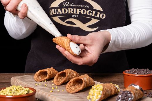 A person piping cream filling into a cannolo shell on a wooden board with bowls of pistachios and chocolate chips.