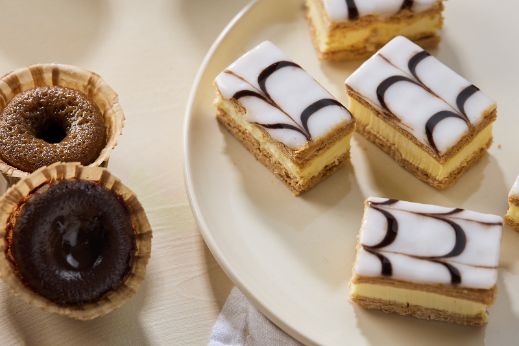 Assortment of pastries including small round tarts and layered iced slices arranged on a light plate and tabletop.