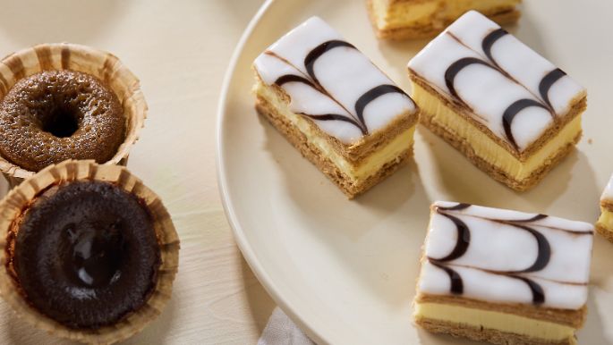 Assortment of pastries including small round tarts and layered iced slices arranged on a light plate and tabletop.
