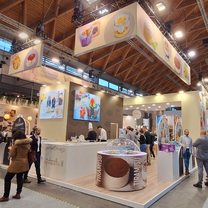 Wide view of the Emmi Desserts stand at SIGEP 2026 showing branded displays, dessert visuals on illuminated panels, a tiramisu-themed counter and visitors walking through the exhibition hall.