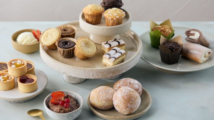 Assorted pastries and mini desserts arranged on ceramic stands and plates, including tarts, muffins, iced slices, rolls and doughnuts.