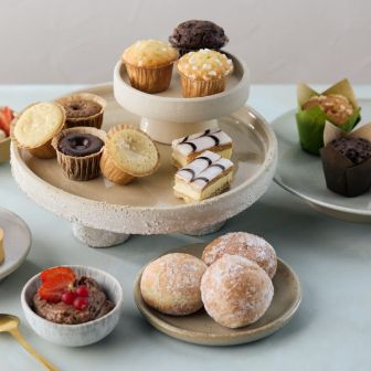 Assorted pastries and mini desserts arranged on ceramic stands and plates, including tarts, muffins, iced slices, rolls and doughnuts.