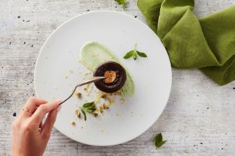 Chocolate lava cake with caramel center served on a white plate with mint, pistachios, and green cream, hand holding a fork
