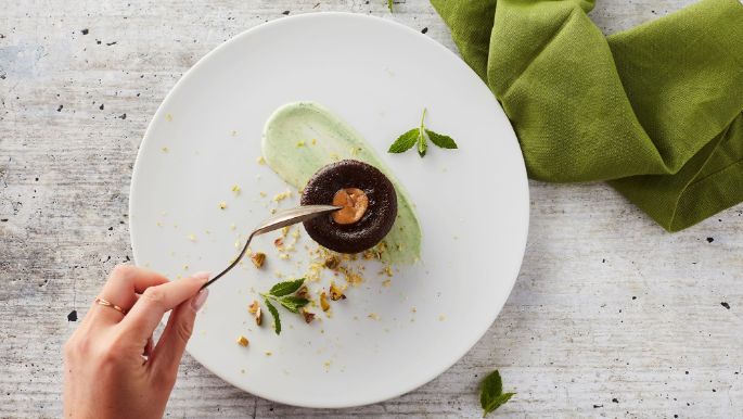 Chocolate lava cake with caramel center served on a white plate with mint, pistachios, and green cream, hand holding a fork
